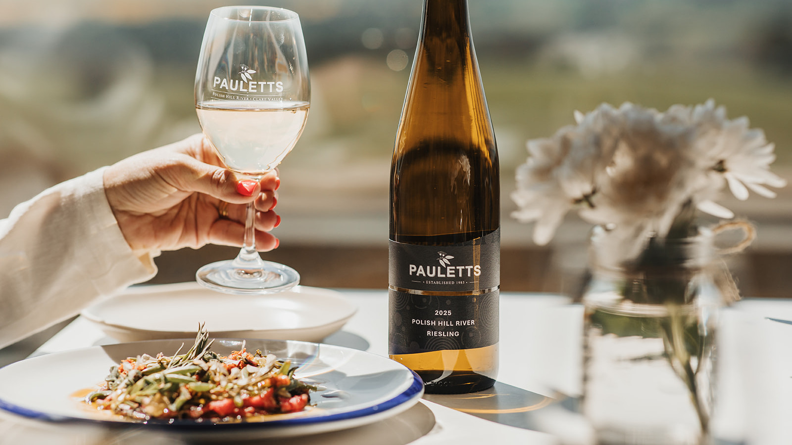 A hand picking up a glass of white wine from a table with a plate of food, a wine bottle and flowers.
