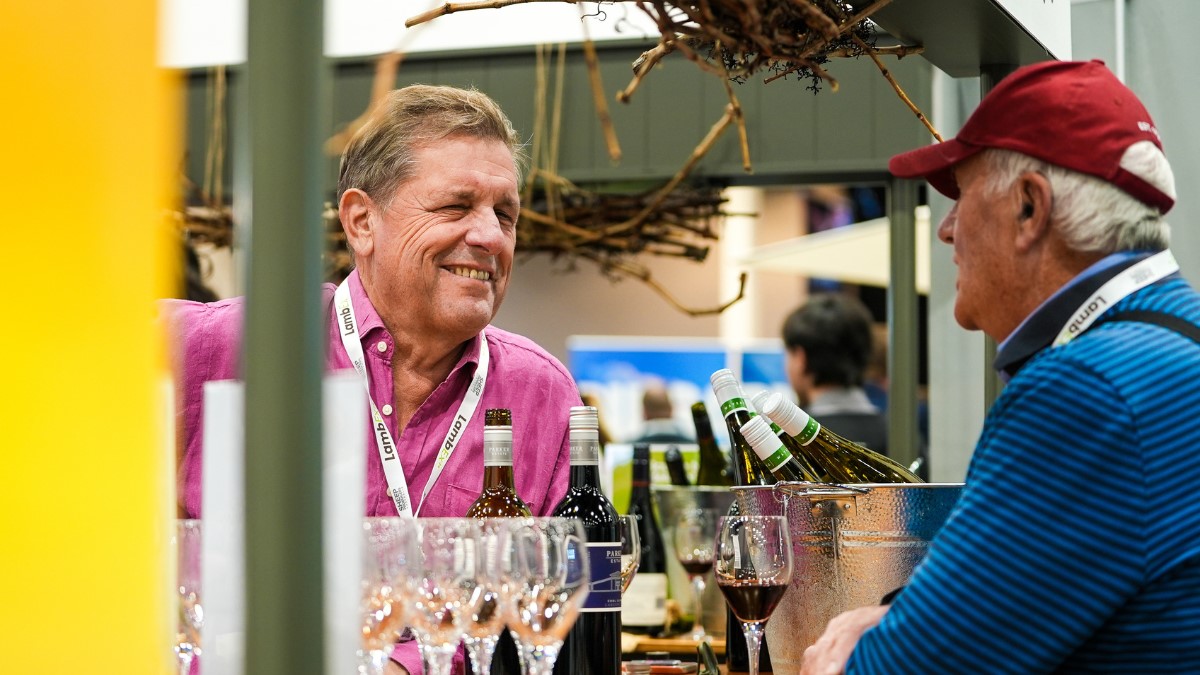 man in a pink shirt behind an outdoor wine tasting booth talking to a man in a red cap 