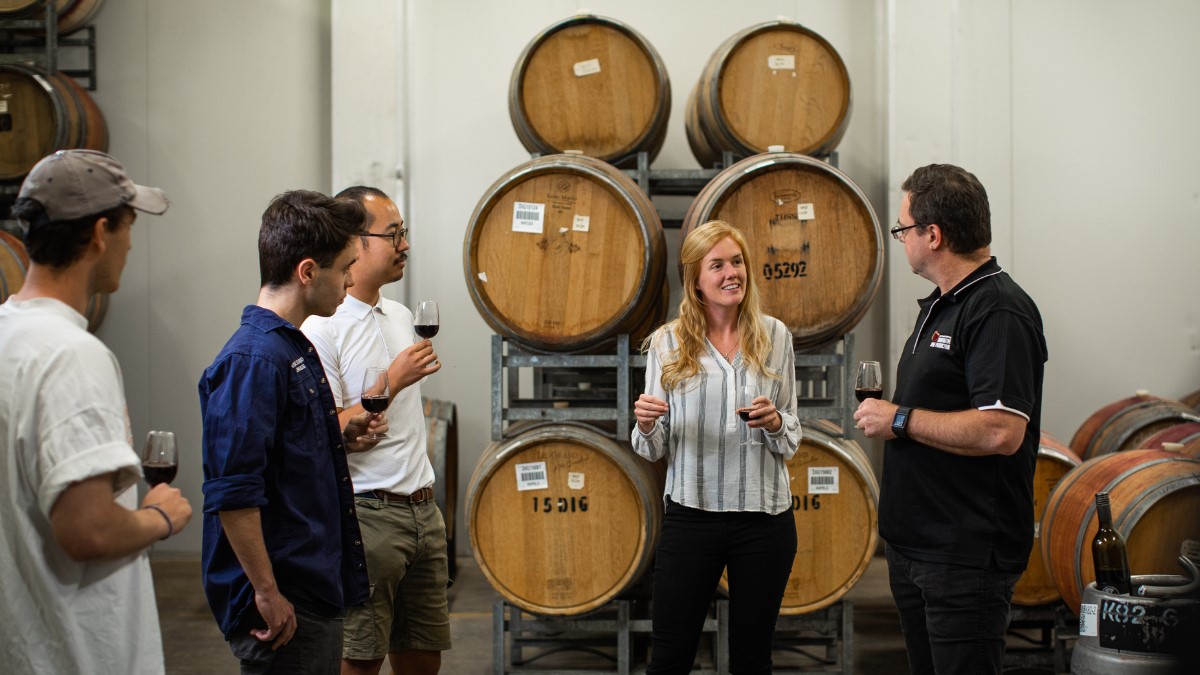 4 men and one woman holding glass of red wine in front of wine barrels