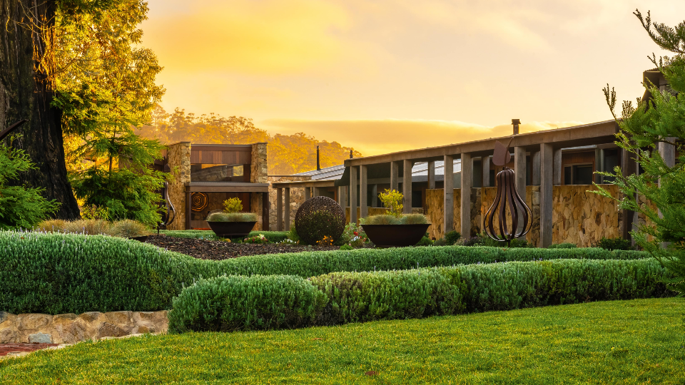 Mount Lofty House and Estate at sunset. Orange skies sit behind an opulent building fronted with green lawns and hedges in the front.